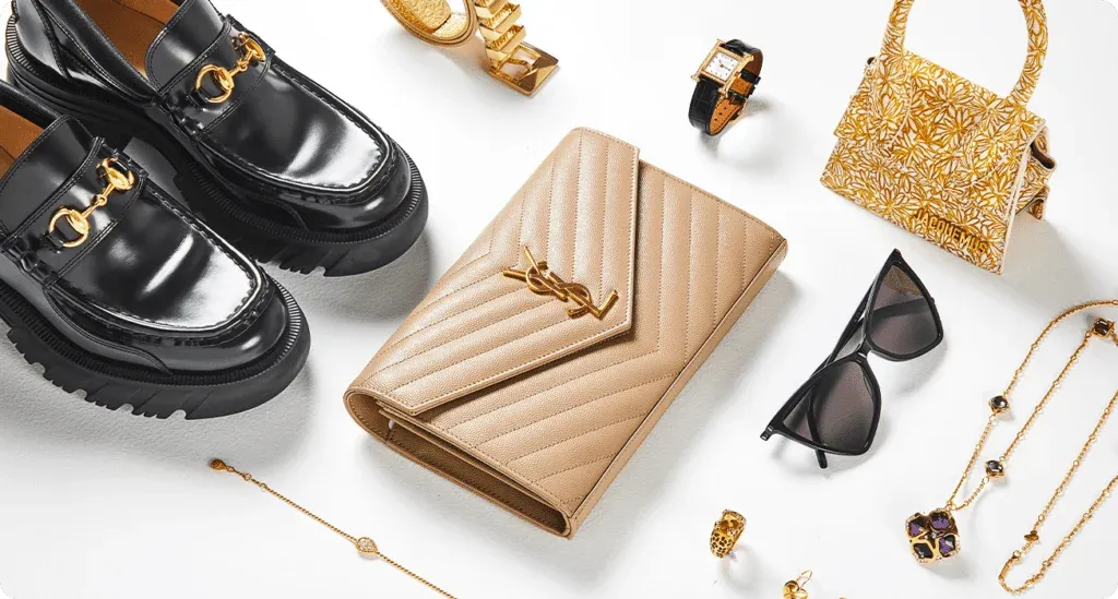 A collection of luxury fashion accessories including black leather loafers with gold detailing, a beige quilted clutch, a gold and floral patterned handbag, black sunglasses, a gold bracelet, a gold watch with a black strap, and gold jewelry with black stones, arranged on a white background.