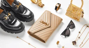 A collection of luxury fashion accessories including black leather loafers with gold detailing, a beige quilted clutch, a gold and floral patterned handbag, black sunglasses, a gold bracelet, a gold watch with a black strap, and gold jewelry with black stones, arranged on a white background.