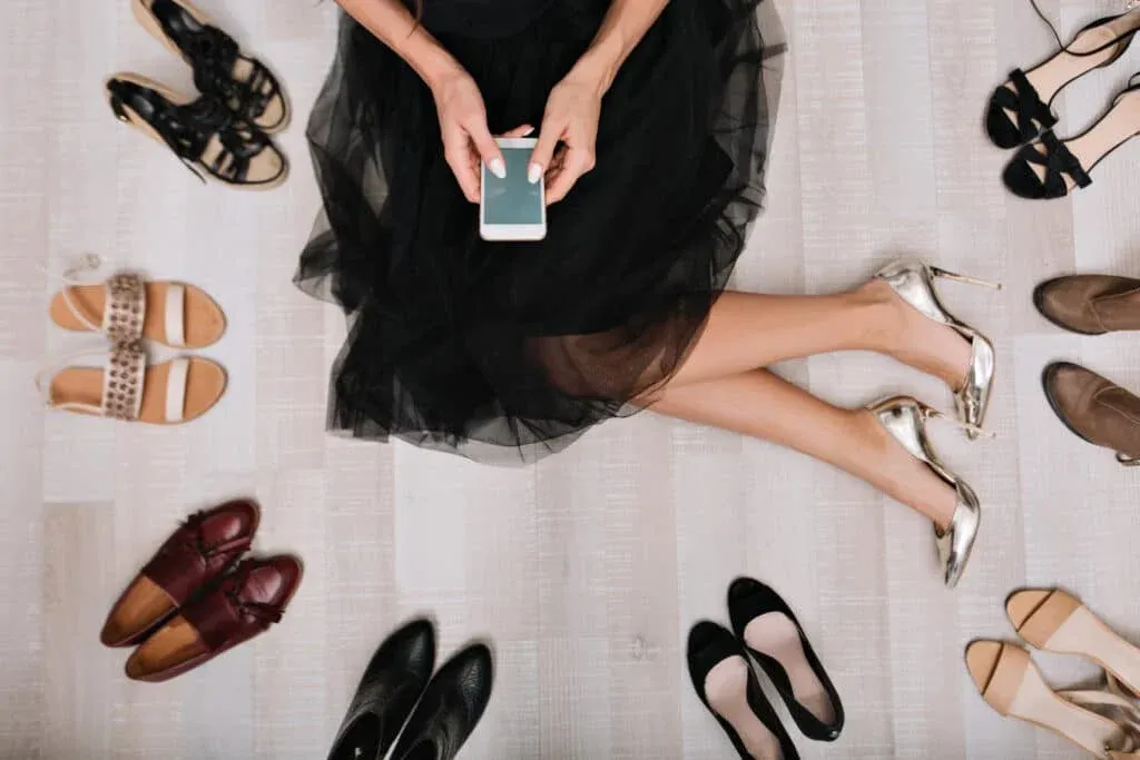 A person sitting on the floor wearing a black dress and silver high heels, holding a smartphone, surrounded by an assortment of different types of shoes including sandals, loafers, and pumps.