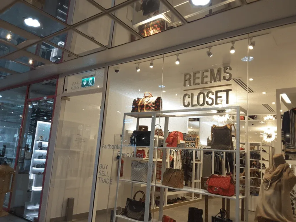 Storefront of 'Reems Closet' with handbags displayed on shelves inside, and a sign indicating the shop deals in pre-owned goods, allowing customers to buy, sell, or trade.