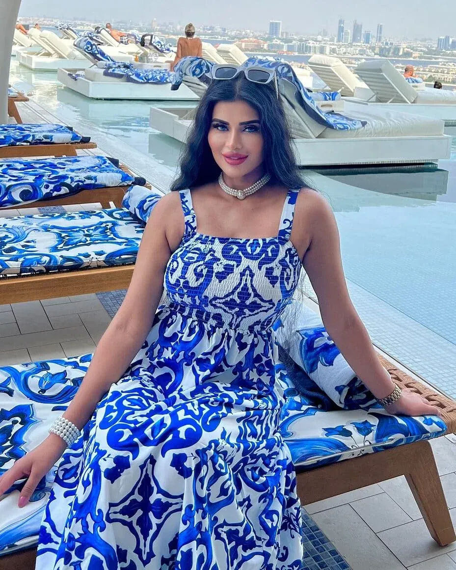 A woman wearing a blue patterned dress sits on a sun lounger by a pool with a cityscape in the background.