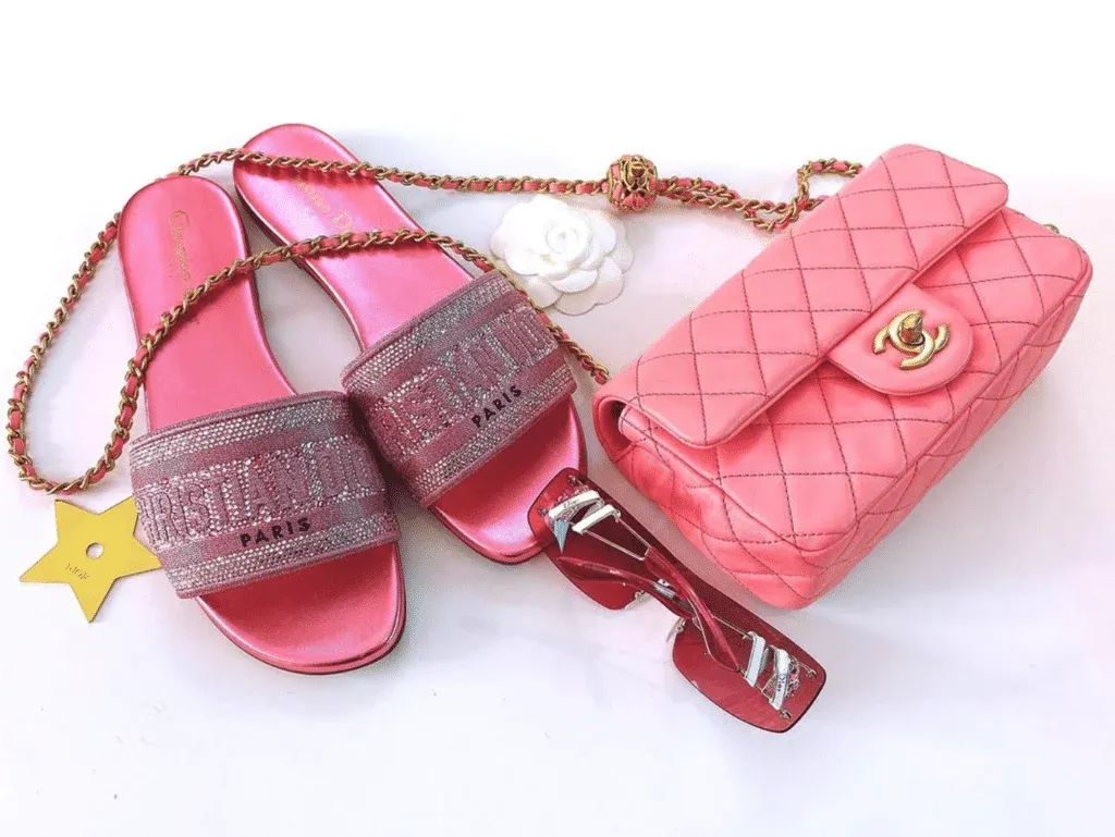 A photo of pink designer sandals, a quilted pink handbag with a gold chain and clasp, and red-tinted sunglasses on a white background.