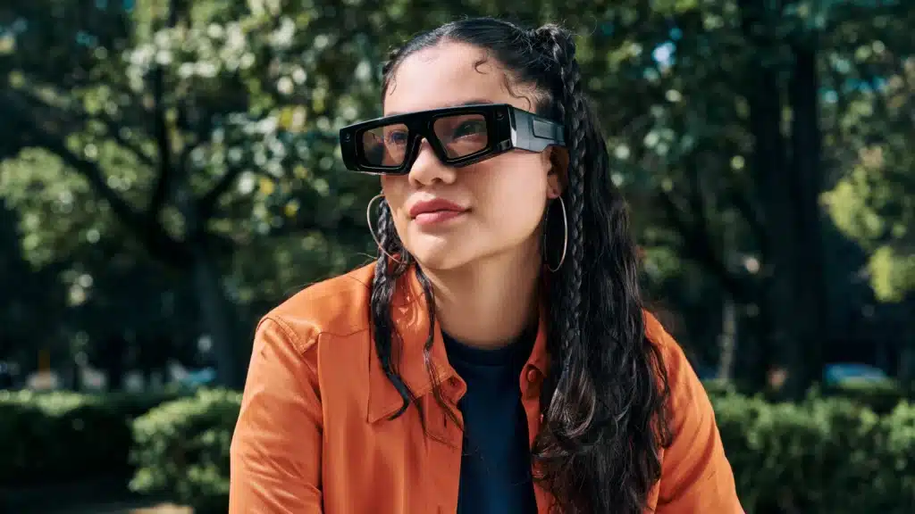 A person wearing large black smart glasses and an orange jacket, sitting outdoors with trees in the background.