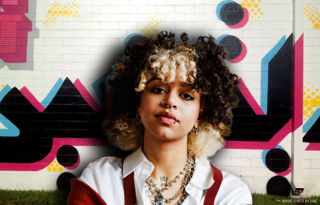 A person with curly hair stands in front of a colorful graffiti wall, featuring geometric shapes in pink, blue, and black.