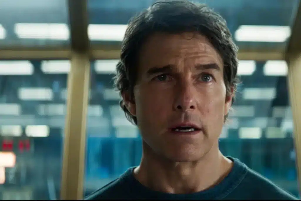 A person with short dark hair, wearing a dark shirt, is indoors with a slightly concerned expression, against a blurry background of a room with windows and lights.
