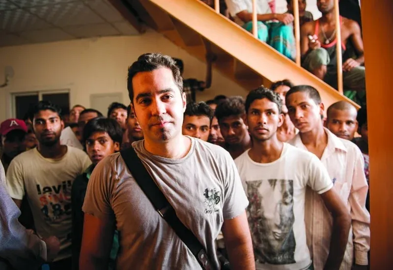 A group of men standing indoors, with one man in the foreground wearing a gray t-shirt and a black strap. The men are gathered under a staircase with orange railings.
