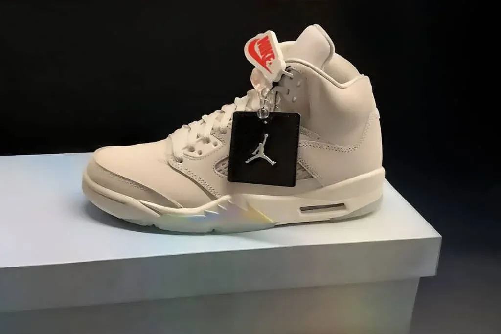 A beige high-top sneaker displayed on a pedestal, featuring a Nike tag and a black tag with a Jumpman logo attached to it.