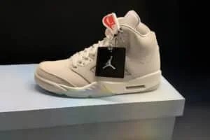 A white high-top basketball sneaker with a visible air unit in the heel and a black tag featuring a silhouetted logo hanging from the laces.