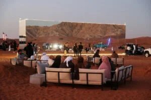 People seated on sofas in a desert area at sunset, facing a large reflective structure that mirrors the surrounding landscape, including cars and mountains.