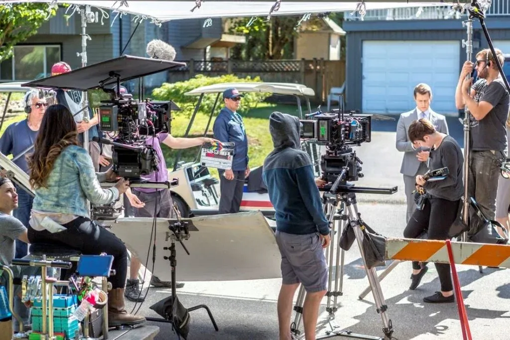 A film crew is on set, filming a scene on a residential street. Several professional cameras are mounted on tripods, and crew members are adjusting equipment and handling a clapperboard. In the background, a male actor in a suit stands next to a woman holding a guitar. The area is shaded by overhead canopies.
