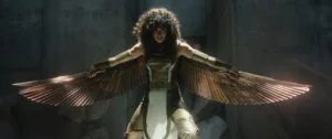 A person wearing an intricately designed costume with metallic wings extended, standing in a dramatic pose in a dimly lit, stone-walled environment.