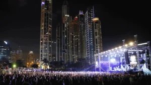 A large outdoor concert at night with a crowd gathered in front of a brightly lit stage, set against a backdrop of illuminated skyscrapers.