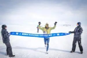 A runner celebrating as they cross the finish line of the World Marathon Challenge 2023 in Antarctica, with two people in winter gear holding the blue race banner in snowy conditions.