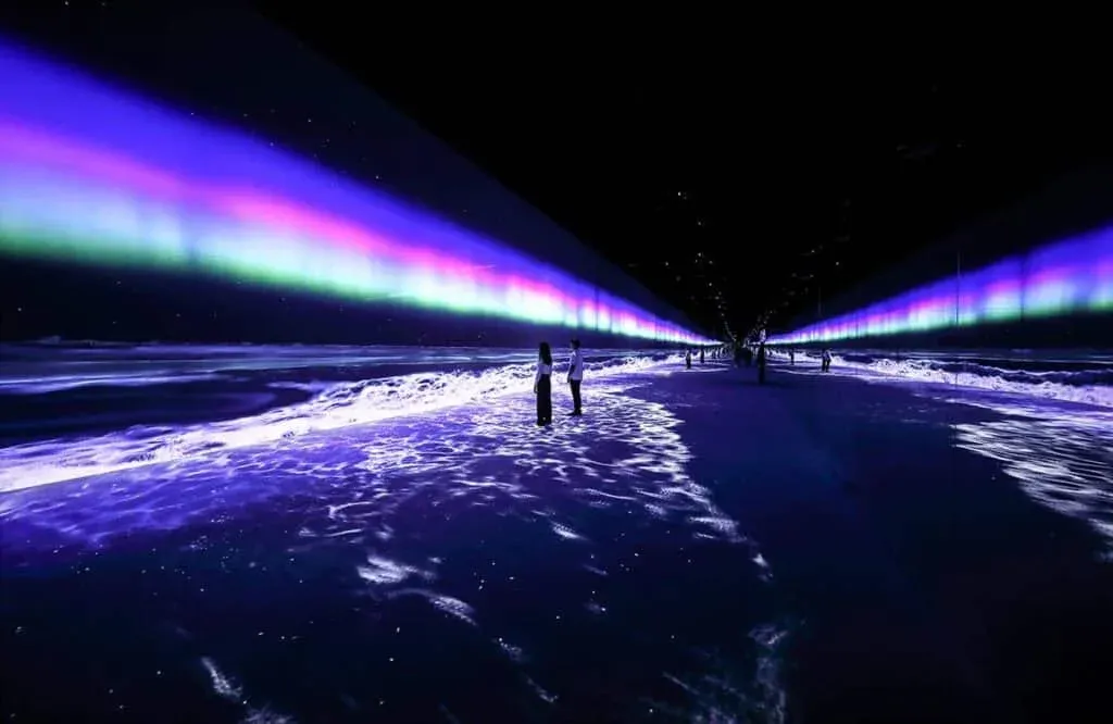 People standing on a reflective floor in a dark room with colorful light beams resembling an aurora, creating an immersive digital art experience.