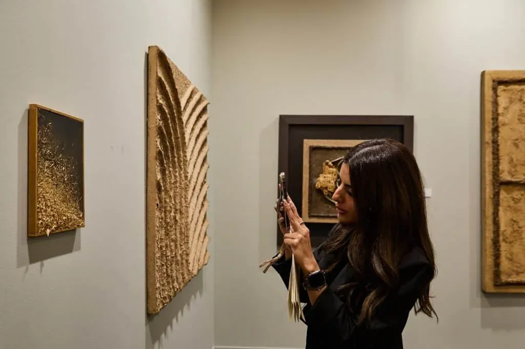 A woman takes a photo with her smartphone of a textured art piece on a gallery wall, surrounded by several abstract artworks in similar earthy tones.