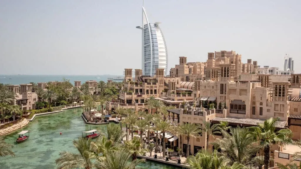 A view of a coastal city with traditional Middle Eastern architecture and a water canal, set against a backdrop of the ocean and a distinctive sail-shaped skyscraper.