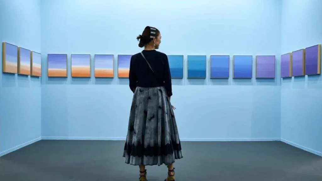 A person standing in a gallery, viewing a series of abstract gradient paintings on light blue walls.