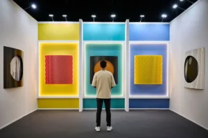 A person stands in an art gallery viewing colorful abstract artworks on the walls, featuring vertical panels with illuminated backgrounds in yellow, turquoise, and blue.