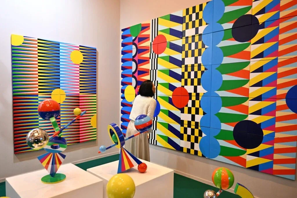 A person observing colorful, geometric art on both the wall and sculptures in a gallery. The wall art features vibrant patterns with shapes like circles and stripes in red, yellow, blue, and black. The foreground shows abstract sculptures with bold colors, spheres, and chrome finishes on pedestal displays.