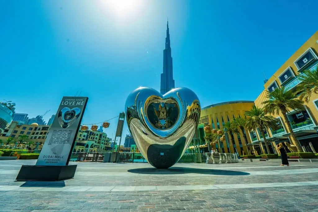 A metallic heart-shaped sculpture reflecting its surroundings stands near the Burj Khalifa, with a bright sunny sky overhead. Palm trees and buildings of Dubai Mall are visible around the sculpture.