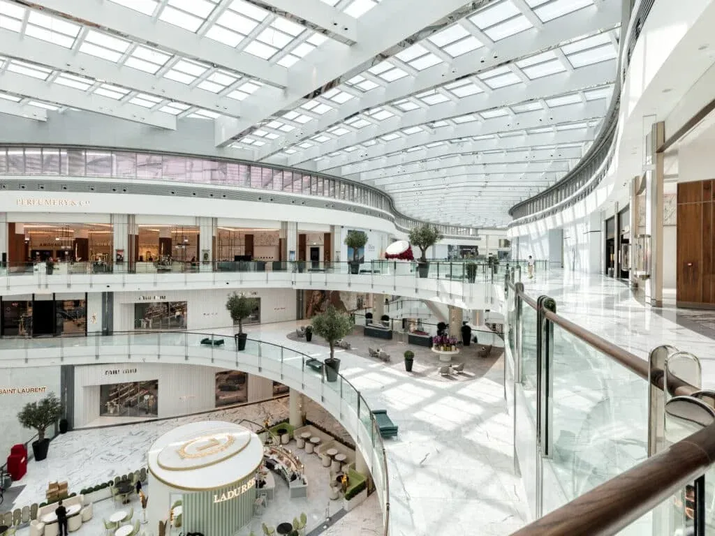 A spacious, modern shopping mall interior with two levels, featuring a glass-domed roof allowing natural light in. The mall has several high-end stores visible, such as Perfumery & Co and Saint Laurent, and a café area on the lower level surrounded by seating. The design includes white marble floors and glass railings.