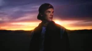 A person wearing a cap looks thoughtfully to the side, standing against a dramatic sunset with a gradient of orange and purple hues in the sky.