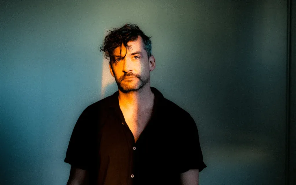 A man with curly hair and a short beard stands against a wall, half-lit by warm sunlight streaming in from the side. He is wearing a dark unbuttoned shirt, and the background is softly shaded in blues and greens.
