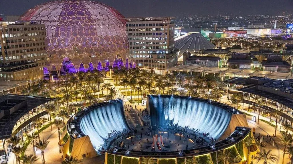 Aerial nighttime view of Expo 2020 site in Dubai, featuring the illuminated Al Wasl Dome and a waterfall installation with surrounding palm trees and buildings.