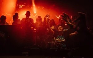 A group of people enjoying a night out at a club, illuminated by warm, red lighting. Some individuals are holding up their phones, while one person in front is raising their arms, creating a lively and joyful atmosphere.