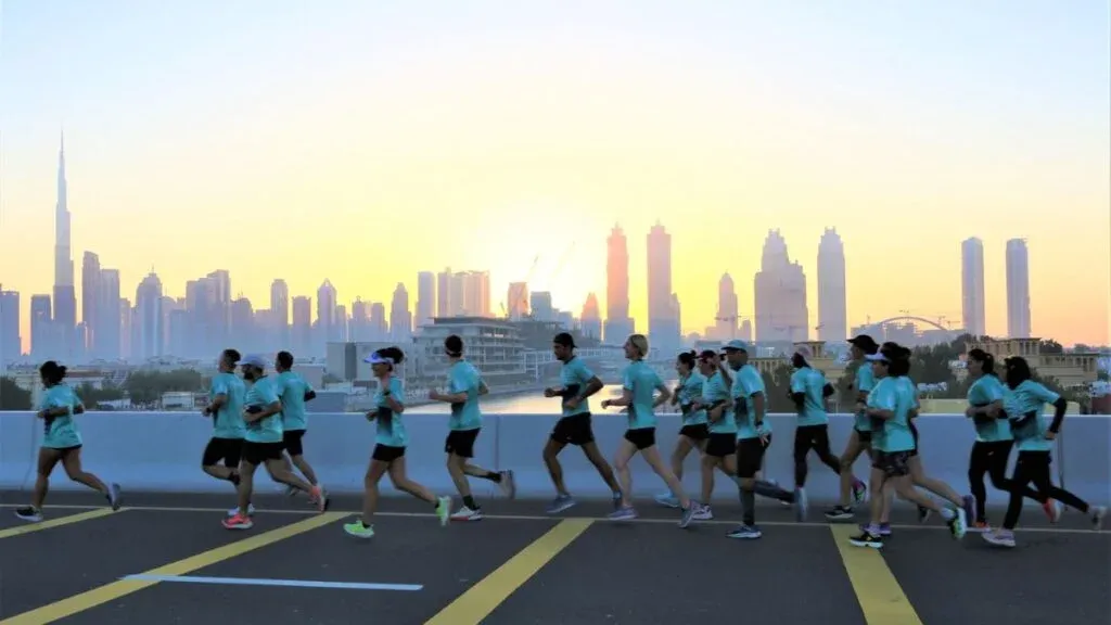A group of people running on a road at sunrise with a city skyline in the background, featuring skyscrapers and a clear sky.