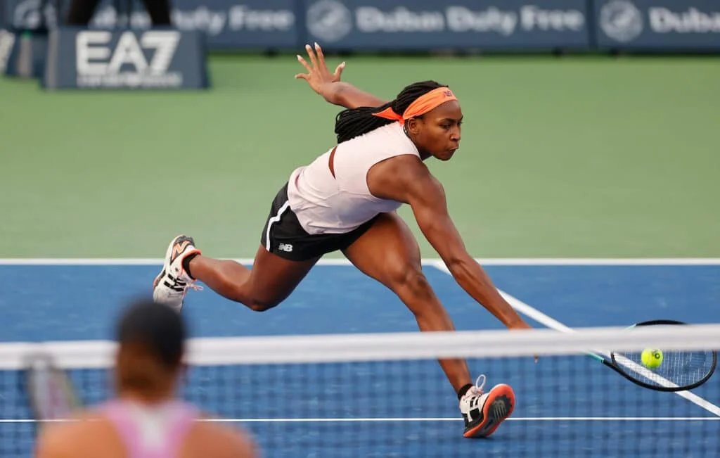 A tennis player reaching for a shot during a match, with one arm extended and eyes focused on the ball, on a blue court.