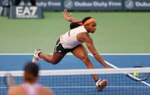 A tennis player reaching for a shot during a match, with one arm extended and eyes focused on the ball, on a blue court.