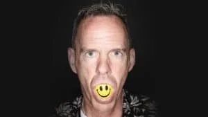 A man with short hair is looking directly at the camera with a yellow smiley face ball in his mouth against a dark background.