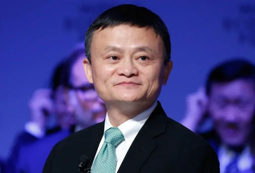 A person in a suit and tie smiling at an event, with a blurred background featuring other attendees.