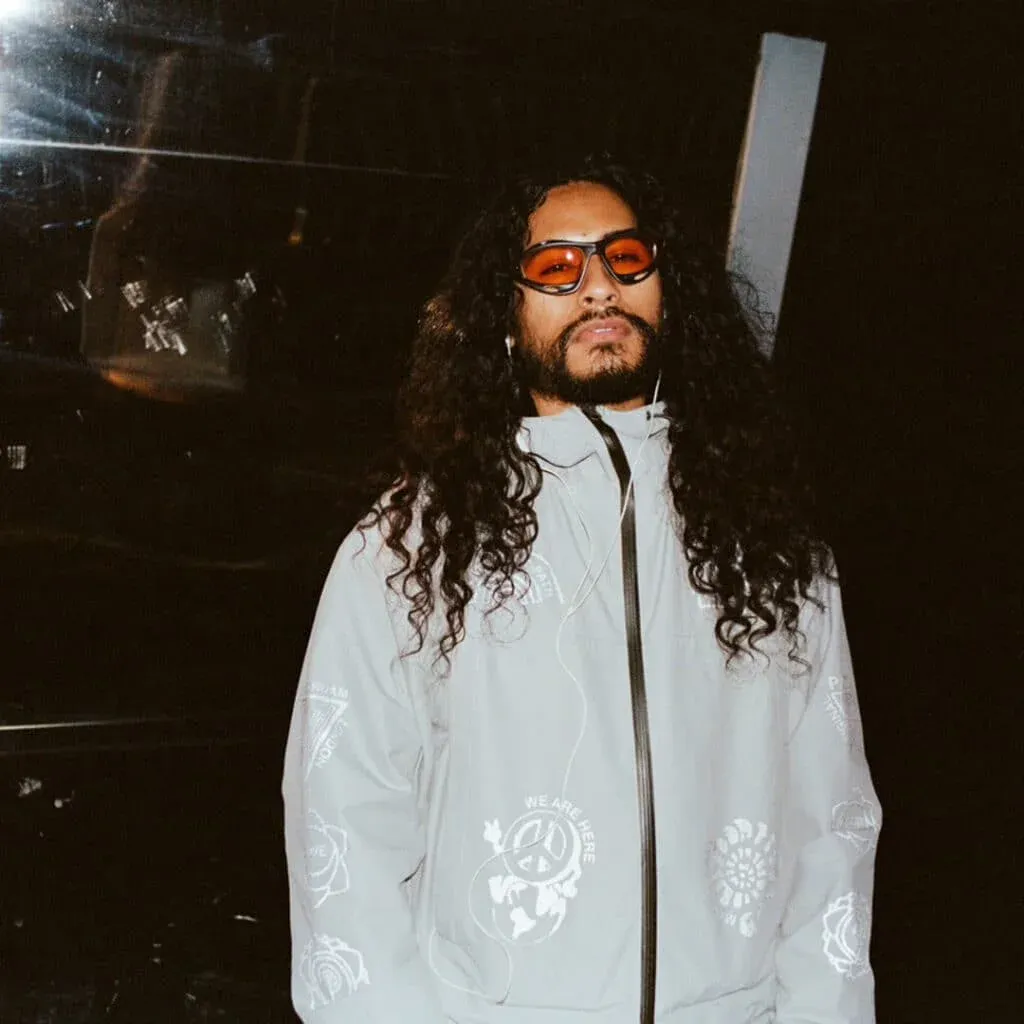 Person with long curly hair and a beard wearing red-tinted sunglasses and a light-colored jacket with graphic designs, standing against a dark background.