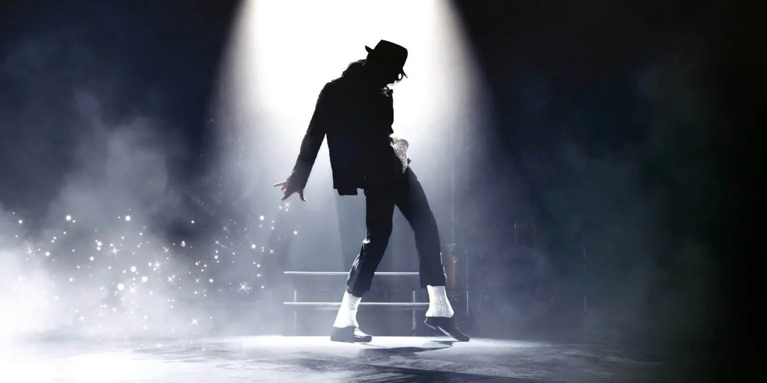 Silhouette of a dancer in a black outfit and hat performing under a spotlight, with light reflecting off glittering socks, surrounded by smoke and sparkles.