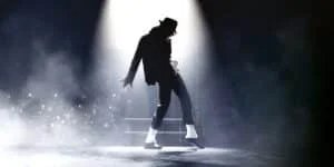 Silhouette of a dancer in a black outfit and hat performing under a spotlight, with light reflecting off glittering socks, surrounded by smoke and sparkles.