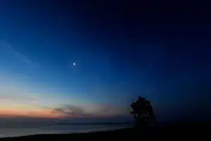 A twilight sky over the ocean with the moon and a few stars visible, along with a silhouette of a lifeguard tower on the right side of the image.