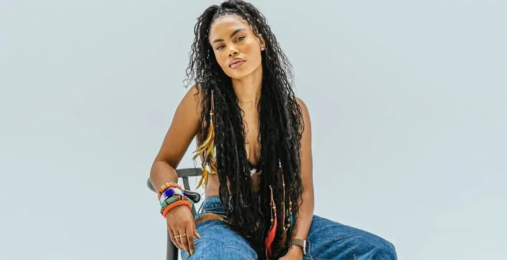 A person with long braided hair wearing a sleeveless top and jeans sits on a chair against a light background. They have colorful bracelets on their wrist and a relaxed expression.