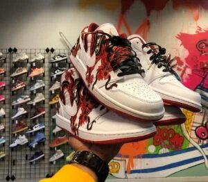 A pair of customized white sneakers with red and black intricate designs, featuring a dragon and floral patterns, held by a person in a store with a colorful wall and various sneakers displayed in the background.