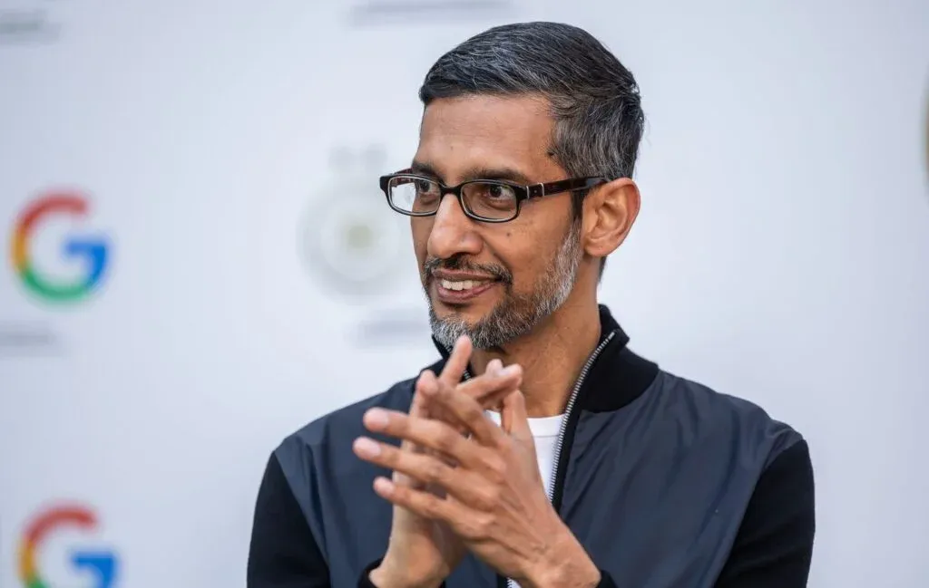 A person with glasses and gray hair is clapping hands, standing in front of a backdrop with a Google logo.