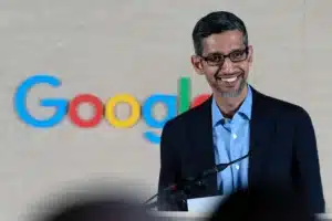 A person in a suit and glasses smiling while speaking at a podium, with a blurred Google logo in the background on a wooden panel.