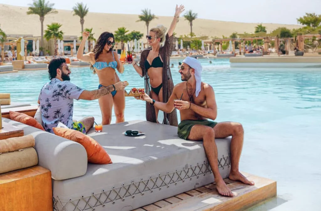 A lively poolside scene featuring four friends enjoying drinks and snacks. Two women in swimsuits and sunglasses are playfully interacting, while two men sit on a lounge, one gesturing with food and the other holding a drink. The pool is surrounded by palm trees and sunny skies, creating a relaxed vacation vibe.
