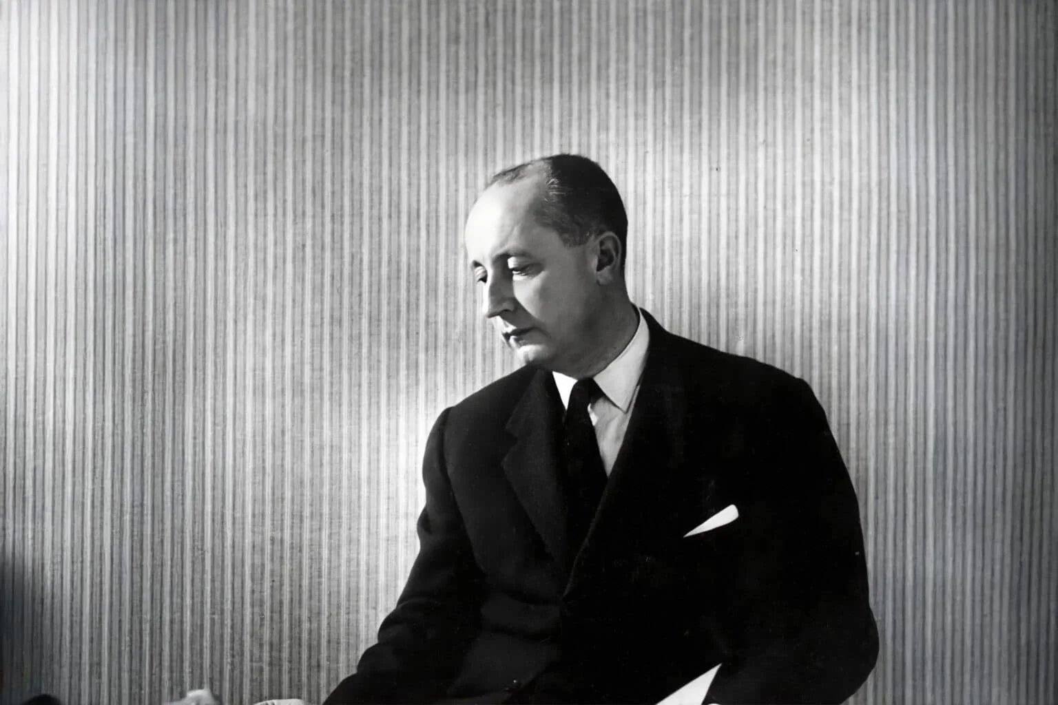 A black and white portrait of a man in a suit, looking downward against a vertically striped background.