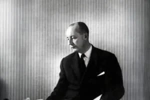 A black and white portrait of a man in a suit, looking downward against a vertically striped background.