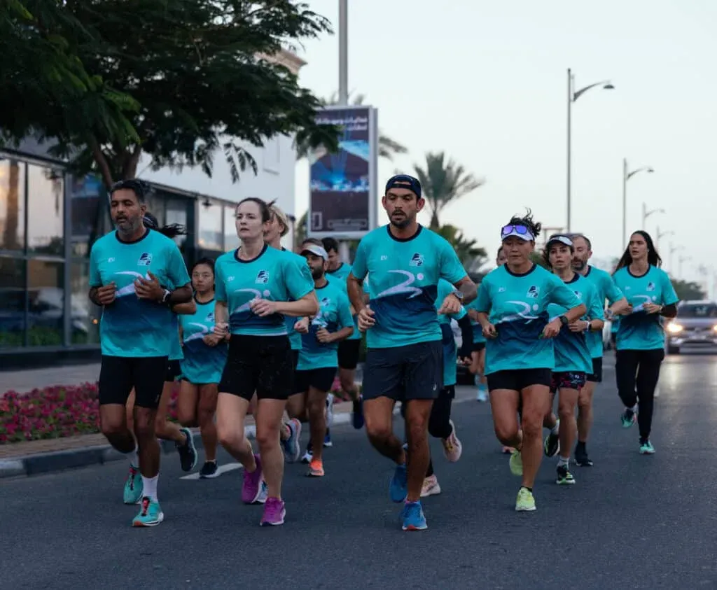 A group of people running together on a street, wearing matching blue athletic shirts and shorts. They appear focused and are participating in a group run or event, with trees and buildings visible in the background.