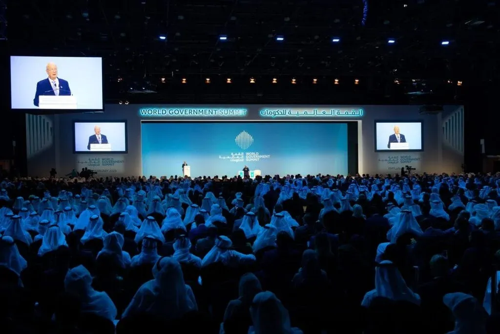 A large audience in traditional attire attending a speaker at the World Government Summit with a blue and white stage backdrop.