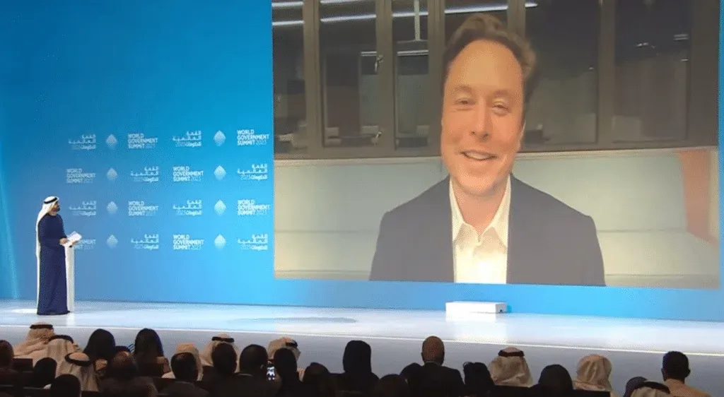 A speaker on stage at the World Government Summit 2023, addressing an audience with a large screen displaying a person in a video call in the background.