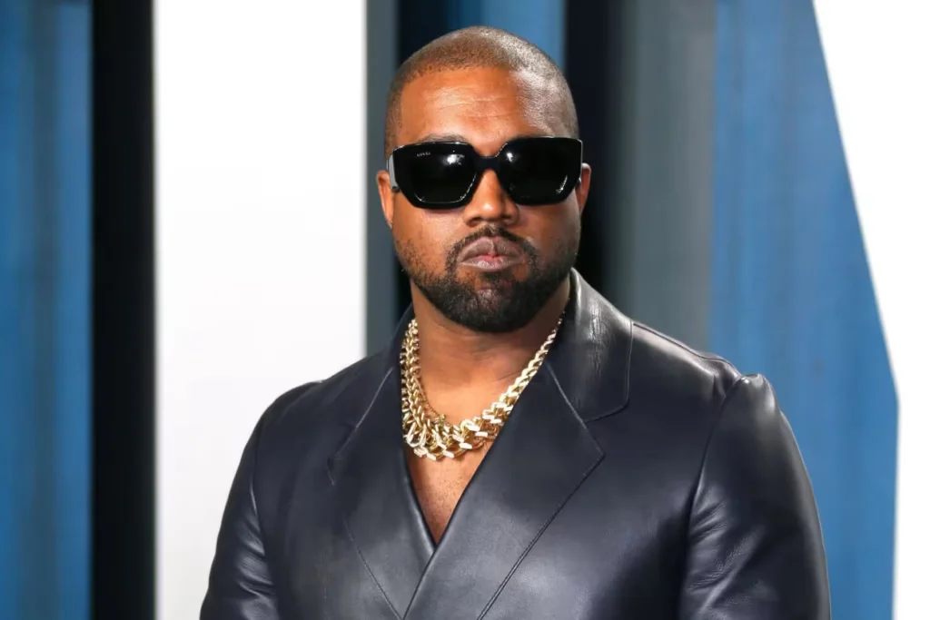 A man wearing dark sunglasses and a black leather jacket with a gold chain necklace in front of a blue and white background.