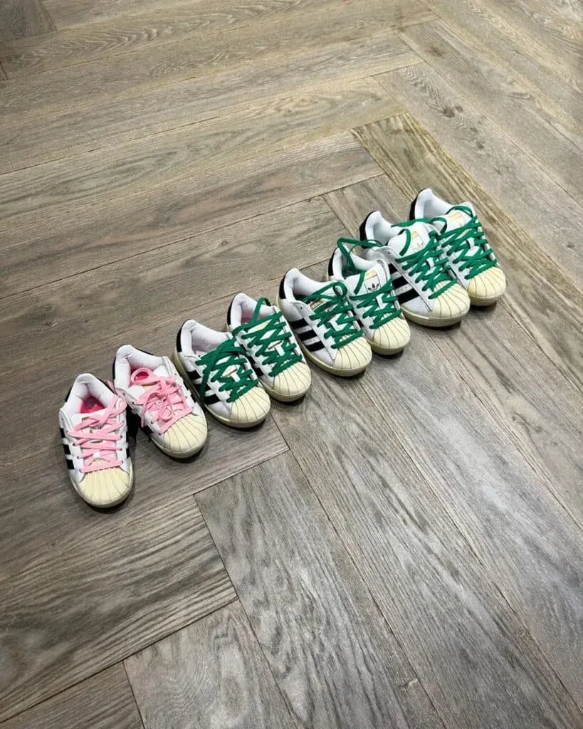 A row of six sneakers in increasing sizes, with white uppers and green or pink laces, displayed on a wooden floor with a herringbone pattern.